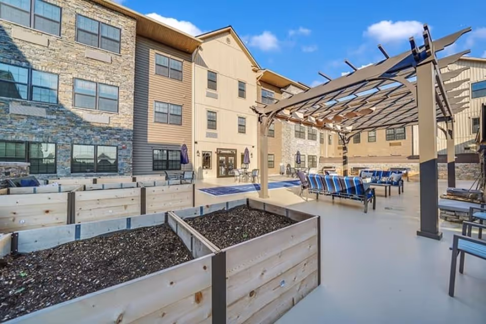 Outdoor patio area at Mercer Hill at Doylestown featuring raised garden beds in the foreground, a pergola with seating underneath, and a multi-story building with stone and siding exterior in the background under a blue sky with some clouds.