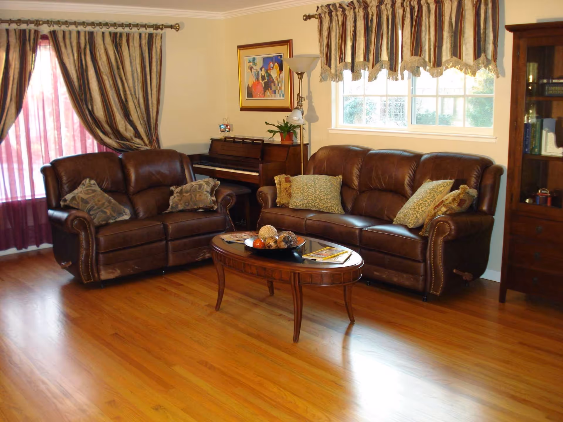 A cozy living room with two brown leather sofas adorned with patterned throw pillows, a wooden coffee table with decorative items and magazines, a piano against the wall, a floor lamp, a framed painting, and windows with striped curtains letting in natural light.