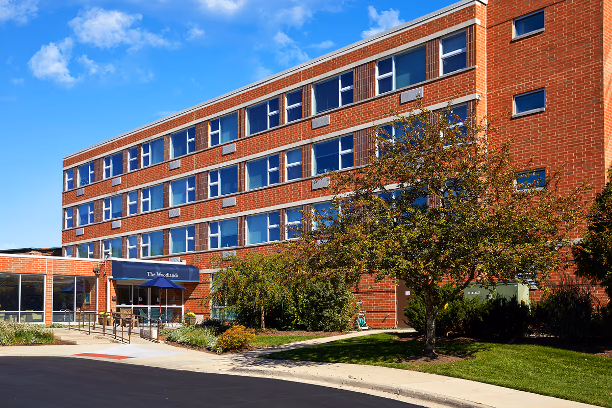 Exterior view of a multi-story brick building with many windows, a blue awning labeled 'The Woodlands' over the entrance, surrounded by green trees and shrubs under a partly cloudy blue sky.