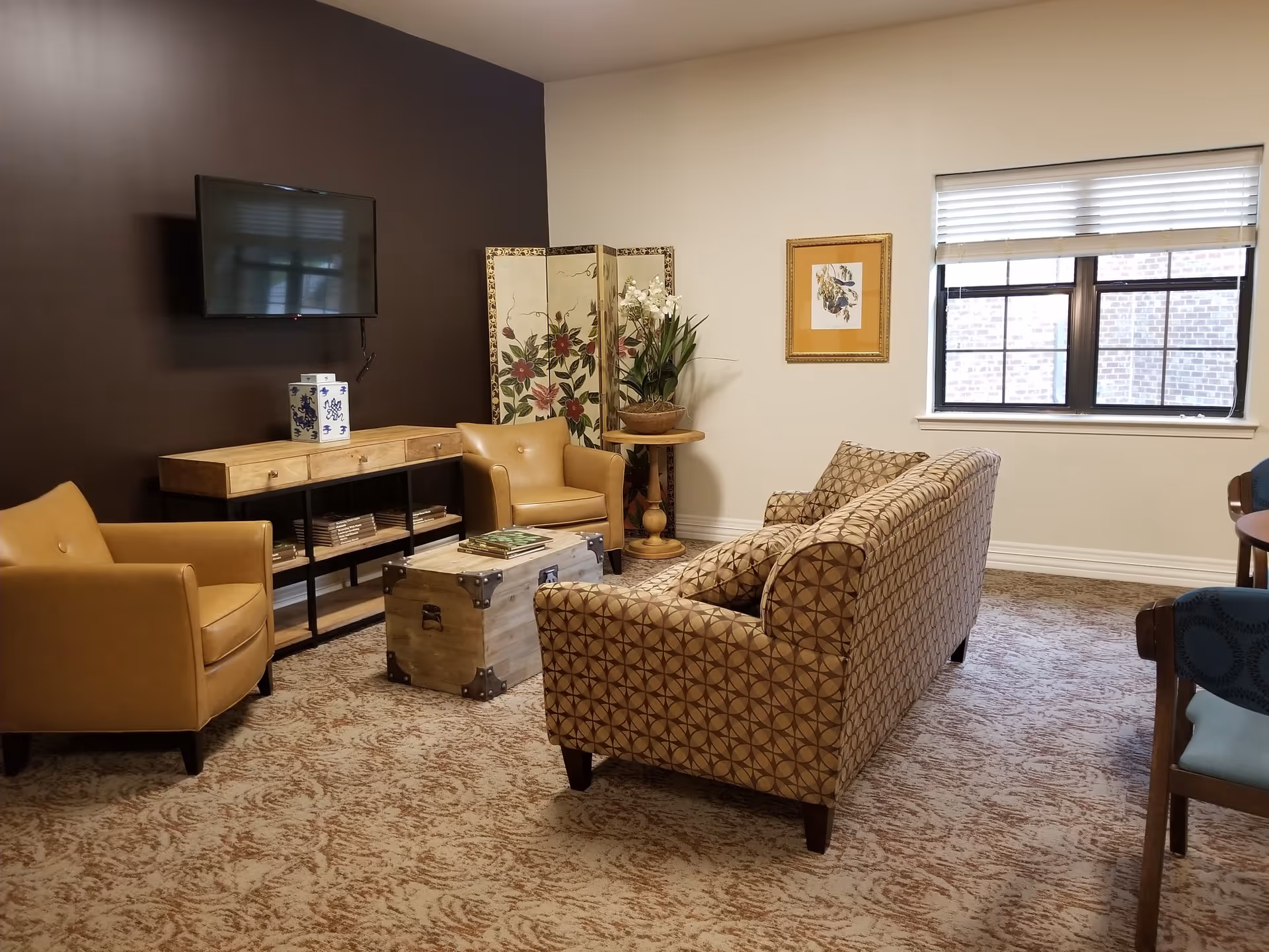 A cozy living room with a patterned sofa, two tan leather armchairs, a wooden coffee table styled like a trunk, a wooden console table with books underneath, a flat-screen TV mounted on a dark accent wall, a decorative folding screen with floral designs, a potted plant on a round side table, a framed picture on the wall, and a window with blinds letting in natural light.