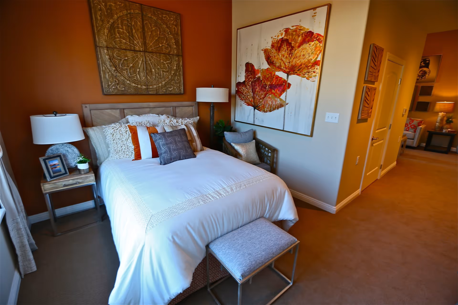 A cozy bedroom with a neatly made bed featuring white and orange pillows. There is a wooden headboard with decorative wall art above it. A small bedside table with a lamp, a framed photo, and a small plant is on the left side of the bed. On the right side, there is a cushioned bench and a large floral painting on the wall. The room has warm-toned walls and carpeted flooring, with a hallway visible in the background.