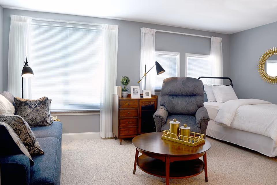 A cozy room with a bed dressed in white linens, a gray recliner chair, a wooden coffee table with decorative items, a blue sofa with patterned pillows, a wooden dresser with framed photos and a small plant, and windows with white curtains and blinds.