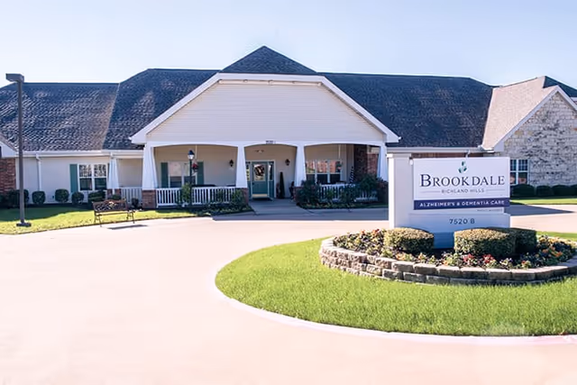 Exterior front view of Brookdale Richland Hills facility with a covered entrance, a bench on the left side, and a landscaped area with a sign displaying the facility name and address.