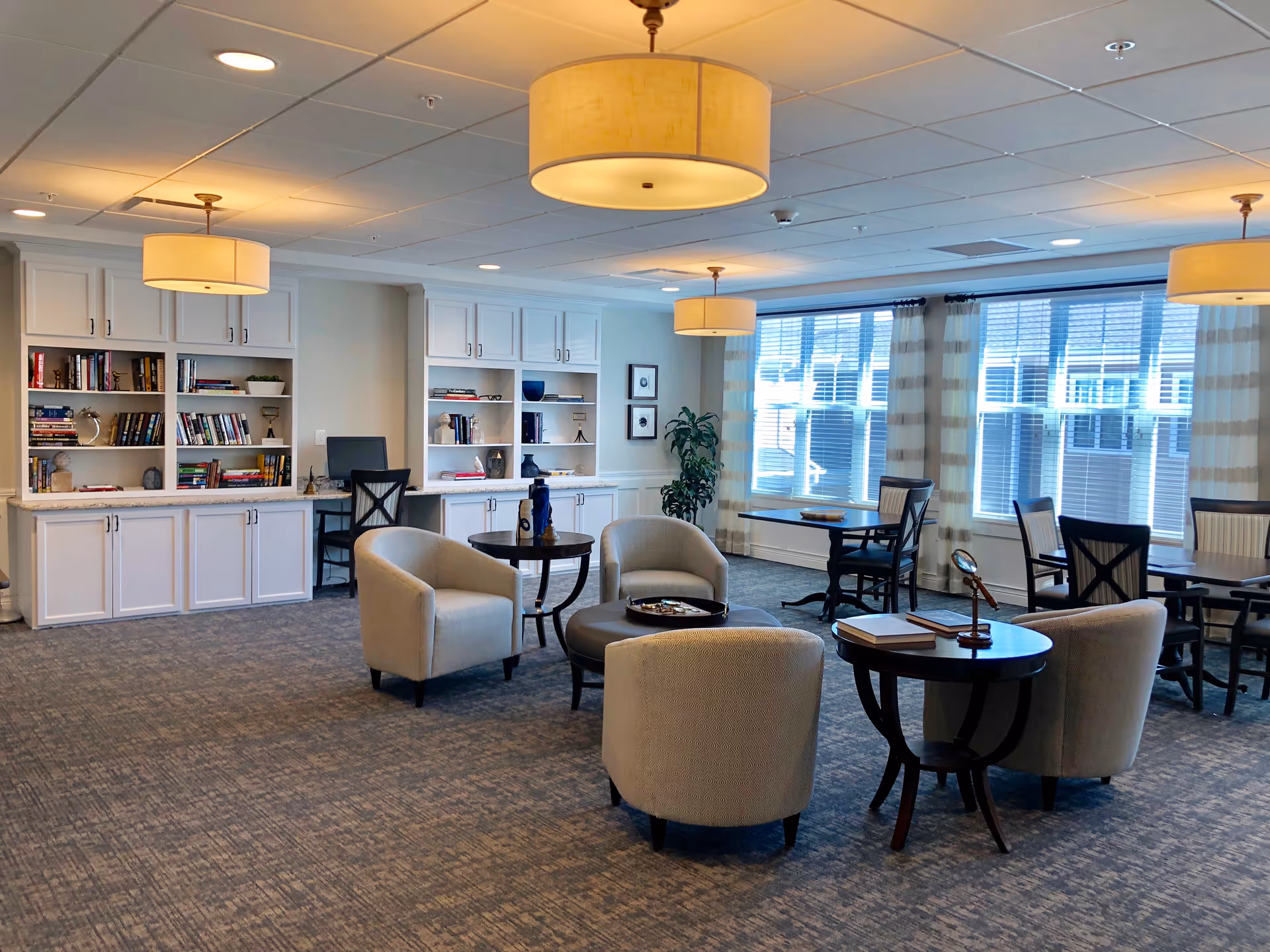 A spacious senior living common area with beige armchairs arranged around small dark wood tables, built-in white bookshelves filled with books and decorative items, large windows with striped curtains allowing natural light, and ceiling-mounted round light fixtures.