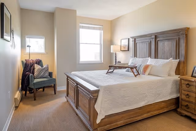 A cozy bedroom with a wooden bed frame and white bedding. There is a wooden nightstand with a lamp and picture frame on the right side of the bed. A small wooden tray is placed on the bed. In the corner near two windows, there is a blue upholstered chair with a blanket and pillow on it. The walls are painted beige and the floor is carpeted.