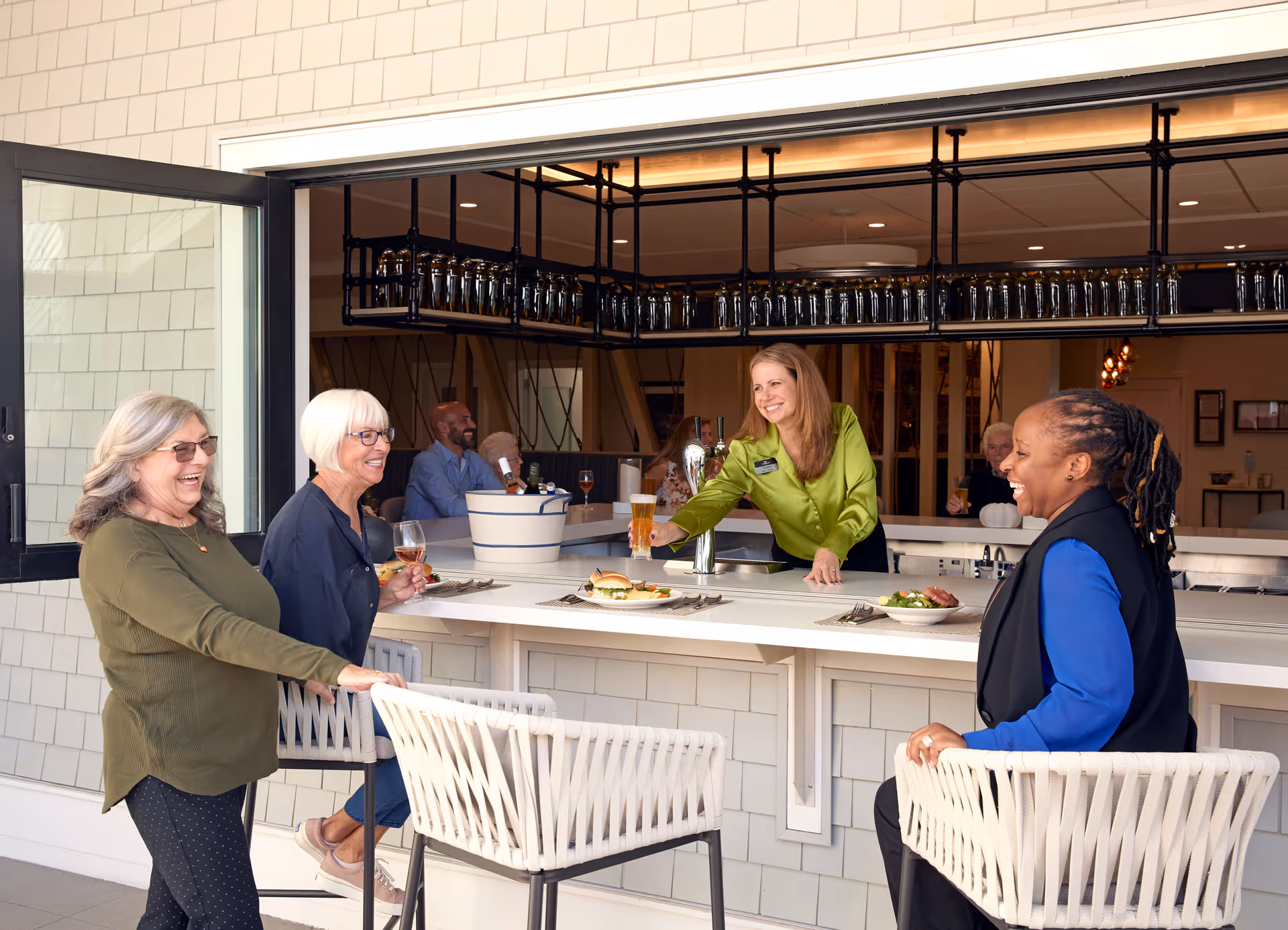 Three women are enjoying drinks and conversation at a bar counter with an open window to an indoor bar area where a bartender in a green blouse is serving a drink. The setting appears bright and welcoming with modern decor and bar stools.