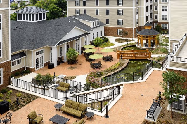 Outdoor courtyard area of a senior living facility featuring multiple seating arrangements with tables and umbrellas, a small pond with a wooden bridge, a gazebo, and surrounding multi-story buildings.