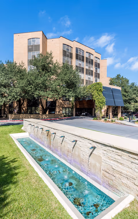 Exterior view of a multi-story beige brick building with large windows, surrounded by trees and greenery. In the foreground, there is a long rectangular water feature with multiple spouts creating small fountains, and a well-maintained lawn beside it. The sky is clear and blue.