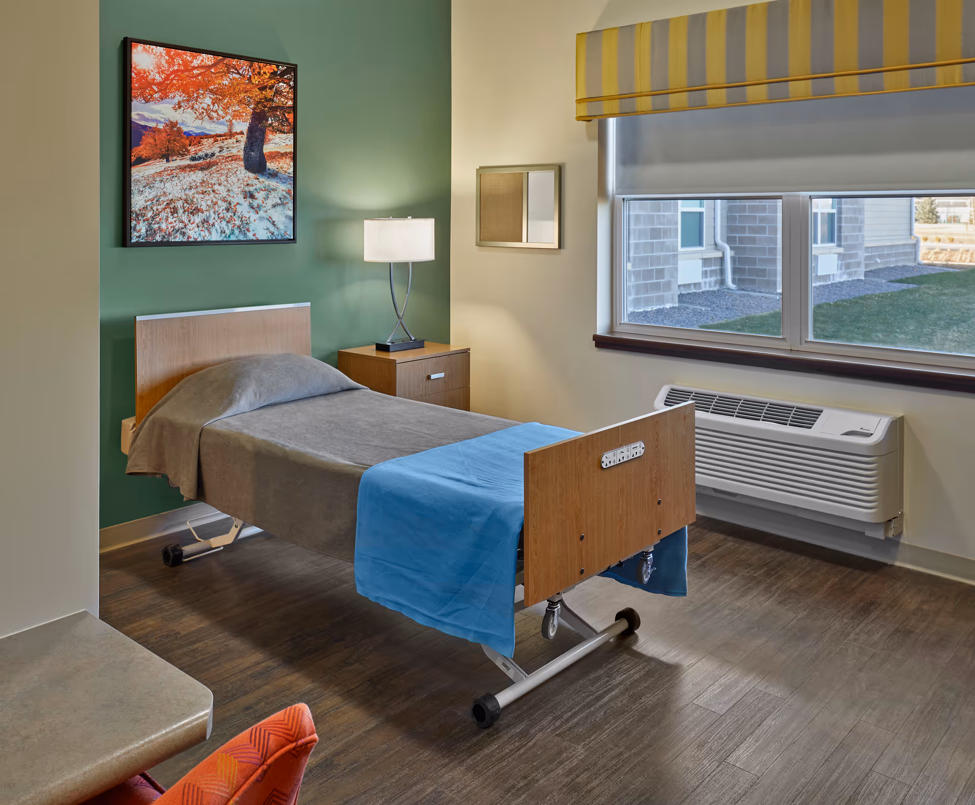 A tidy single-bed resident bedroom with a bedside table and lamp, green accent wall with artwork, window and air conditioning unit.