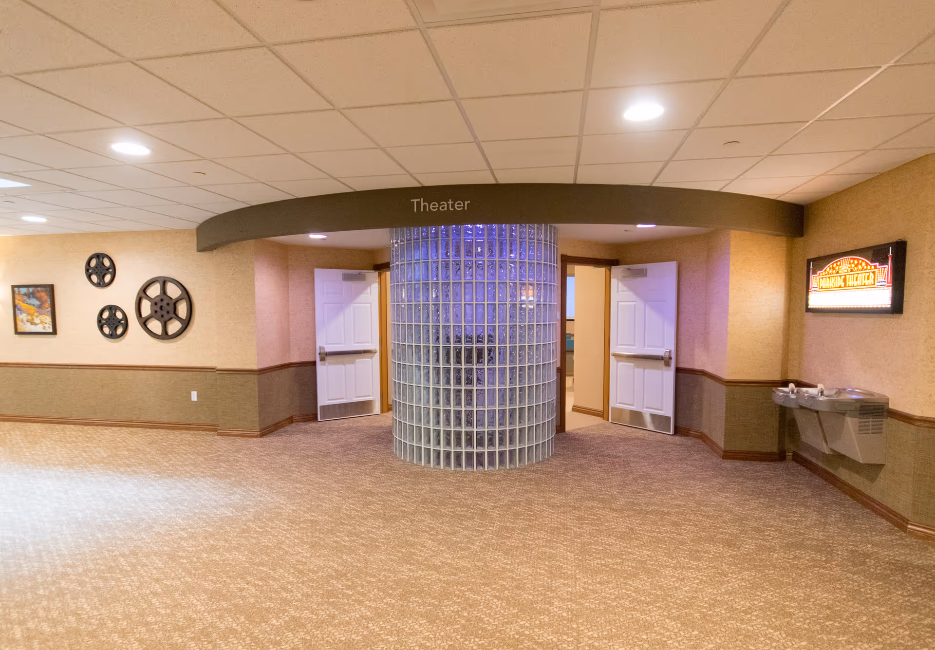 Interior hallway area in a senior living facility with beige carpet and walls. A curved glass block wall is centered with two white double doors on either side leading to a theater room. Above the glass block wall is a curved sign that reads 'Theater'. On the left wall are decorative film reels and a framed picture. On the right wall is a water fountain and a lit sign that says 'Marquee Theater'.