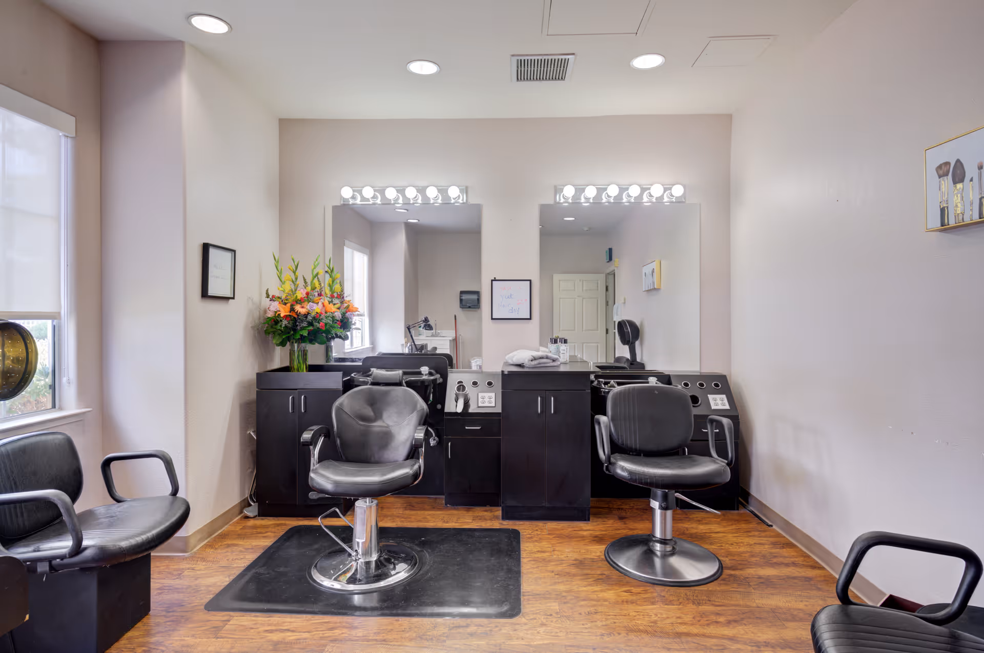 Interior view of a hair salon area with two black salon chairs in front of large mirrors with bright vanity lights. There are black cabinets and hair washing stations behind the chairs. The room has wooden flooring, a window with a roller shade on the left, and framed pictures on the walls.