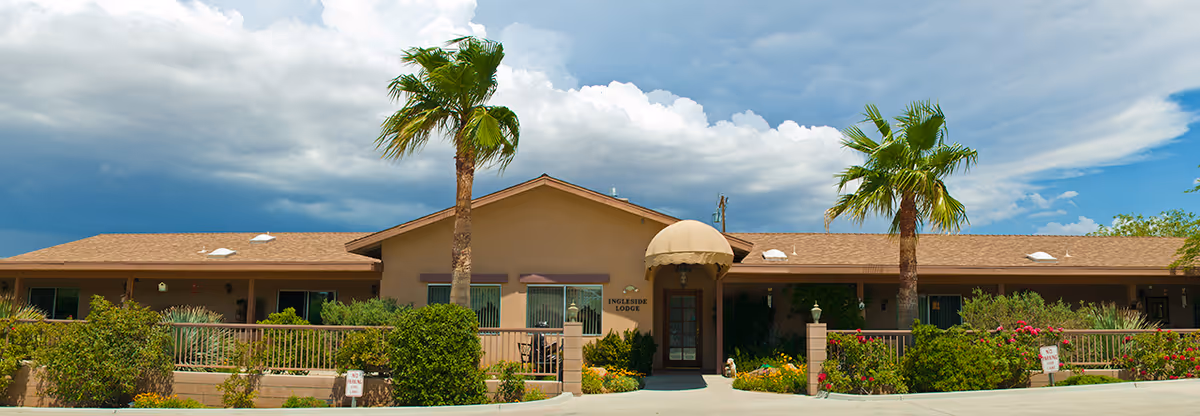 Front exterior view of Ingleside Lodge Assisted Living facility with a single-story building, two tall palm trees, a covered entrance, and landscaped bushes and flowers under a partly cloudy sky.