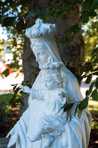 White statue of a crowned woman holding a crowned child, surrounded by green leaves and trees in an outdoor setting.