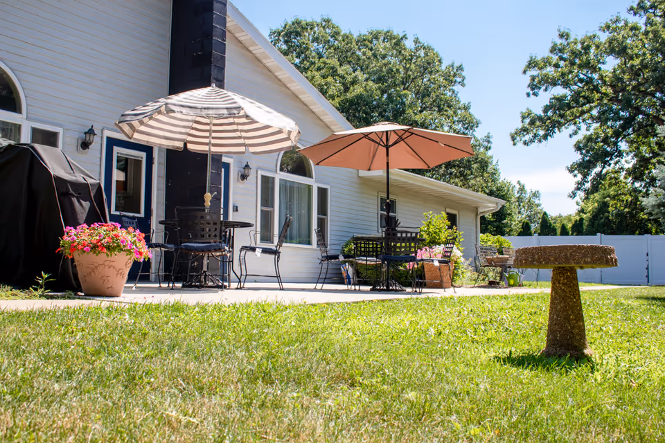 Outdoor patio area of a senior living facility with two tables, each shaded by a large umbrella. There are several chairs around the tables, a barbecue grill covered with a black cover, potted flowers, and a birdbath on a grassy lawn. The building has white siding and multiple windows, with trees and a white fence in the background under a clear blue sky.