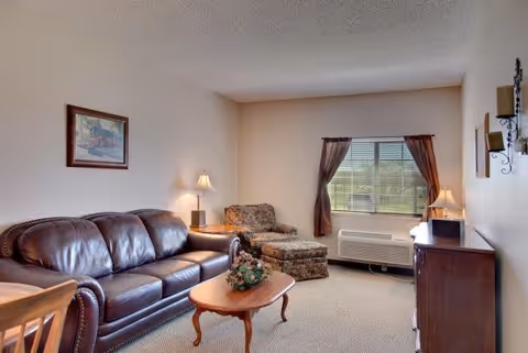 A cozy living room with a dark brown leather sofa, a floral upholstered armchair with matching ottoman, a wooden coffee table with a floral centerpiece, a wooden side table with a lamp, a window with brown curtains, and a wooden cabinet with a lamp on top. The walls are light-colored and there is a framed picture hanging above the sofa.