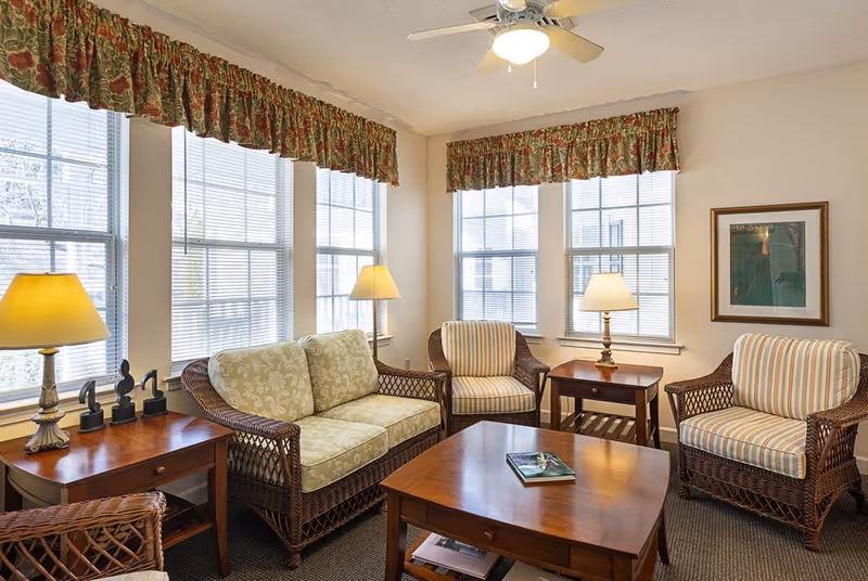 Bright common living room with wicker sofa and armchairs, wooden coffee and side tables, lamps, and multiple windows with floral valances.