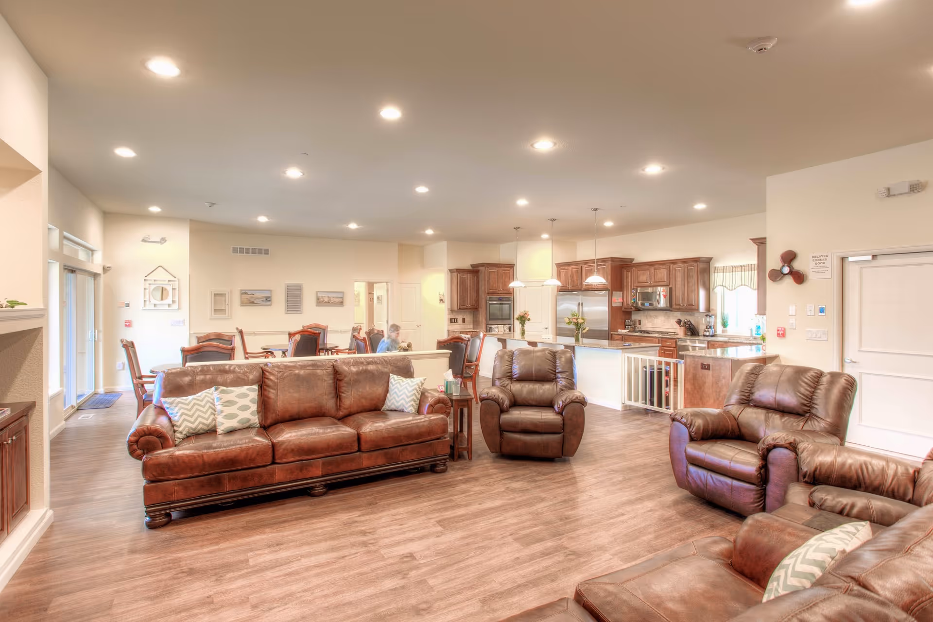 Spacious and well-lit common area in an assisted living facility featuring brown leather sofas and armchairs arranged on wood flooring. In the background, there is a kitchen with wooden cabinets, stainless steel appliances, and pendant lighting. A dining area with tables and chairs is also visible, with a few people seated and engaged in conversation.