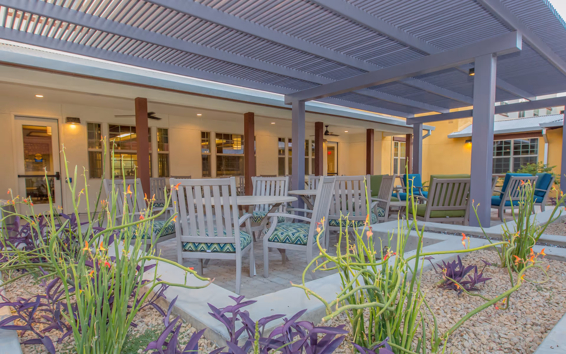Outdoor patio area at The Ranch Estates of Tucson featuring multiple tables and chairs with patterned cushions under a pergola. Surrounding the seating area are desert plants and gravel landscaping, with the building's exterior and windows visible in the background.