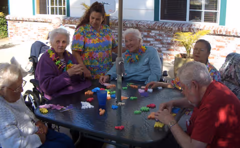 A group of elderly people and a caregiver sitting around a glass outdoor table playing a colorful puzzle game. They are wearing leis and appear to be enjoying a sunny day in a garden or patio area with brick walls and windows in the background.