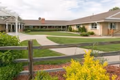 Single-story building with a brown roof and beige walls surrounding a well-maintained green lawn and paved walkways, viewed from behind a wooden fence with yellow flowering plants in the foreground under a partly cloudy sky.