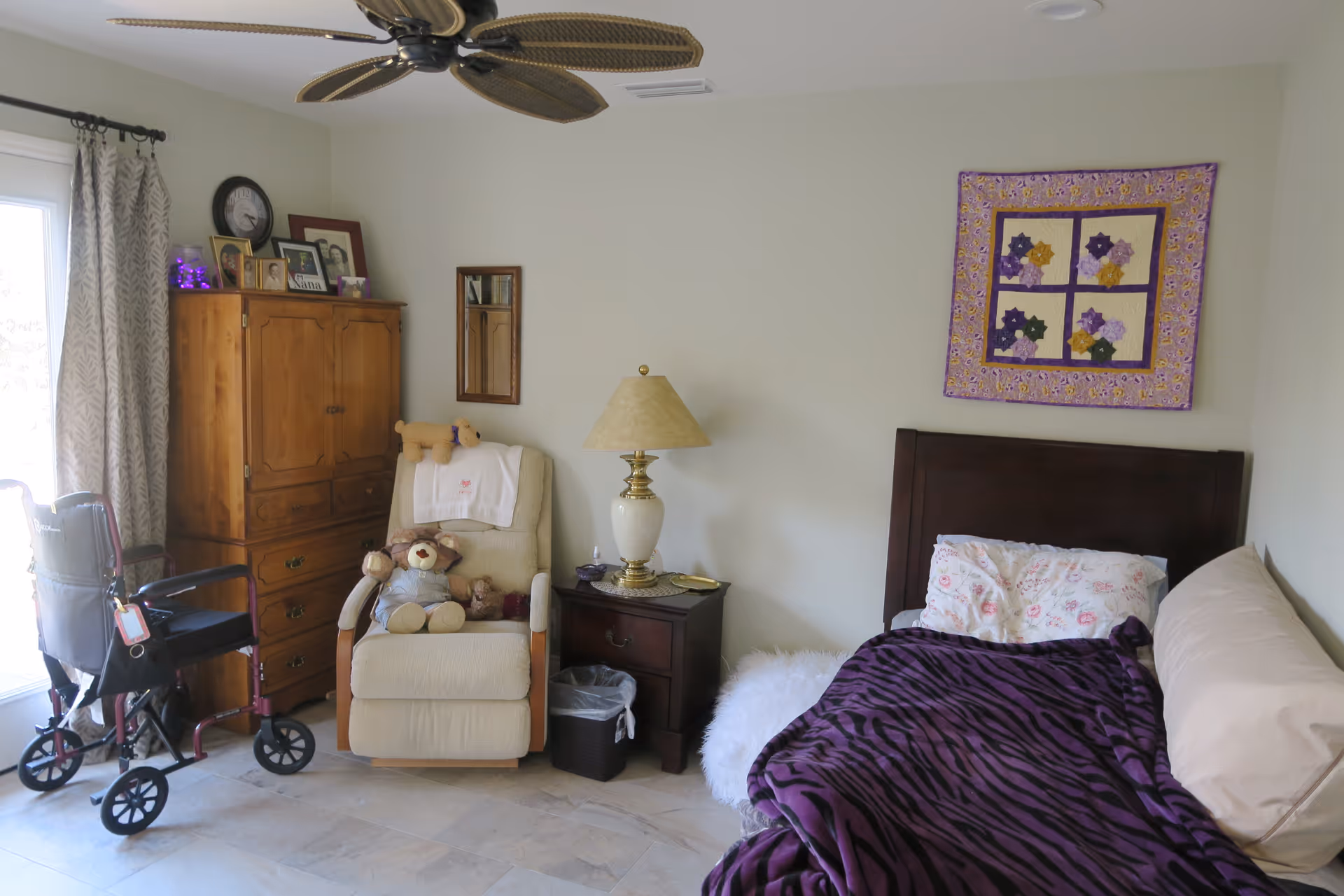 A cozy senior living bedroom with a single bed covered in a purple zebra-striped blanket and floral pillow. Next to the bed is a nightstand with a lamp and a small trash bin underneath. A beige recliner chair with stuffed teddy bears sits beside a wooden cabinet adorned with framed photos and a clock. A walker is positioned near a window with patterned curtains. A ceiling fan with leaf-shaped blades is visible above, and a colorful quilt hangs on the wall above the bed.