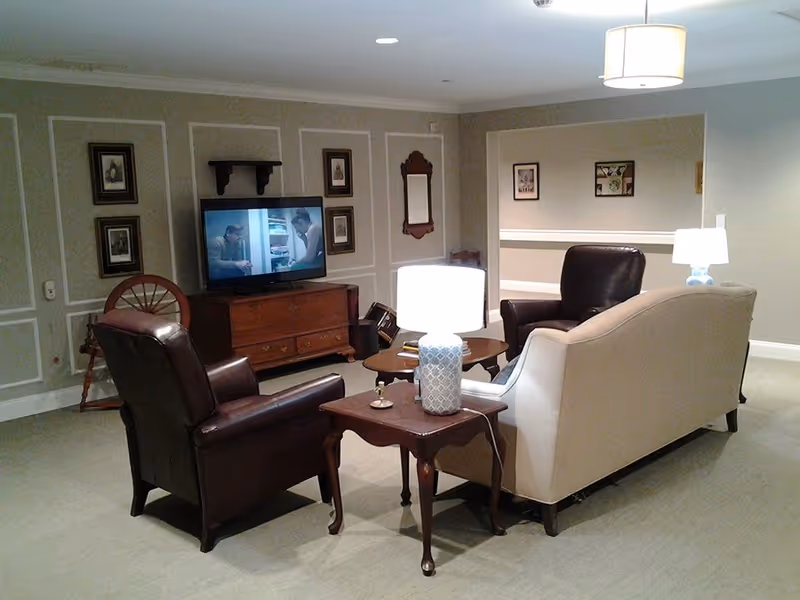 A cozy living room area with two dark leather armchairs and a beige sofa arranged around a wooden coffee table and side table with a lamp. A flat-screen TV is mounted on a wooden cabinet against the wall, which is decorated with framed pictures and a mirror. The room has soft lighting and neutral-colored walls with decorative molding.
