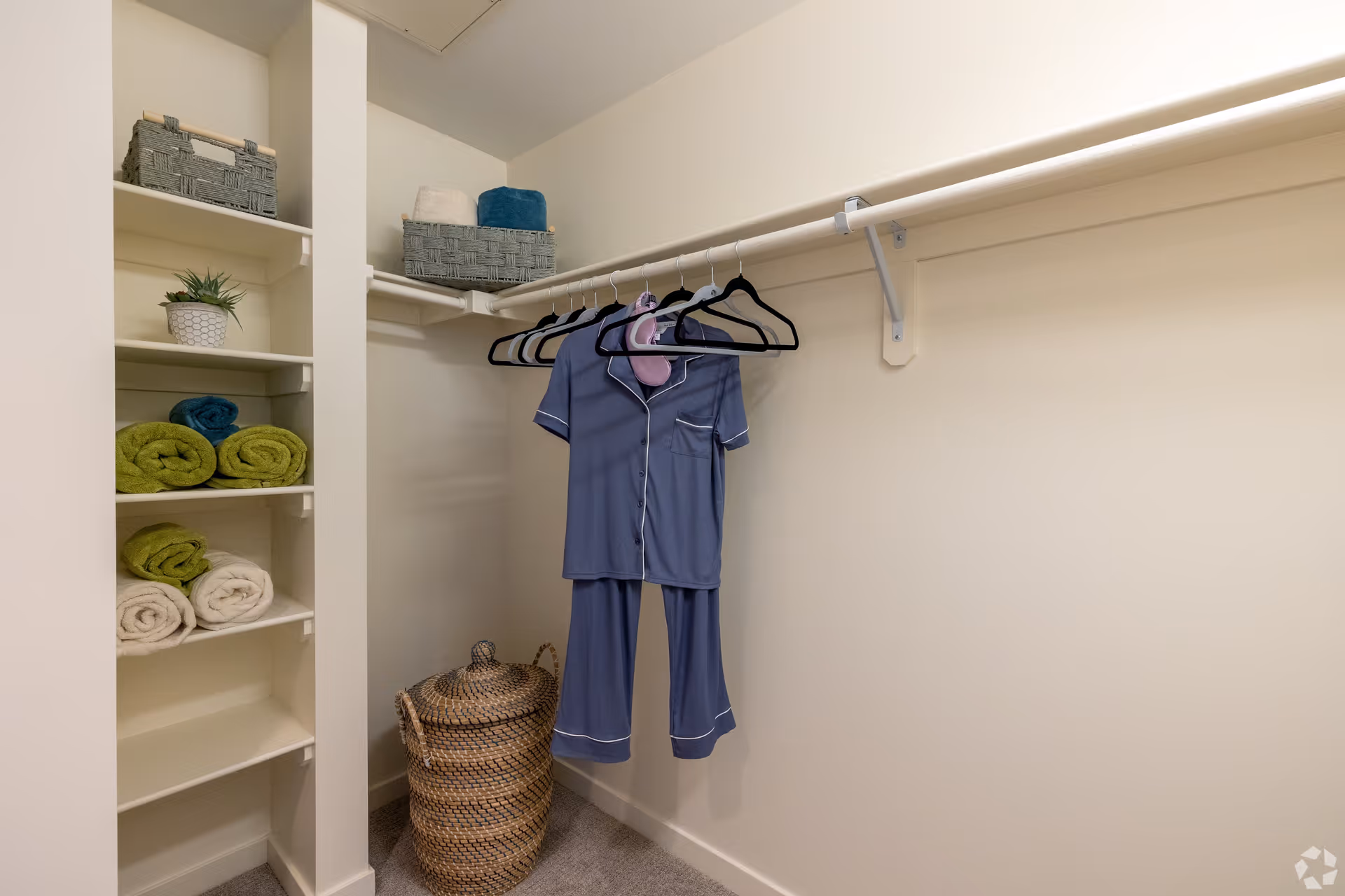 A walk-in closet with a hanging rod holding several empty hangers and a blue pajama set. Shelves on the left side hold rolled towels in green, blue, and white, a small potted plant, and two gray storage baskets. A woven basket with a lid is on the floor beneath the hanging clothes.