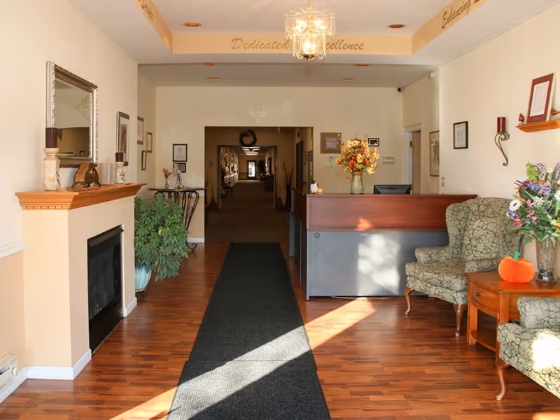 Reception area of DeSmet Retirement Community with a wooden floor, a black runner rug, a fireplace with a mirror above it on the left, a reception desk with a flower vase on top, and two patterned armchairs with a small wooden table holding a flower arrangement on the right. The hallway extends into the background with framed pictures on the walls.