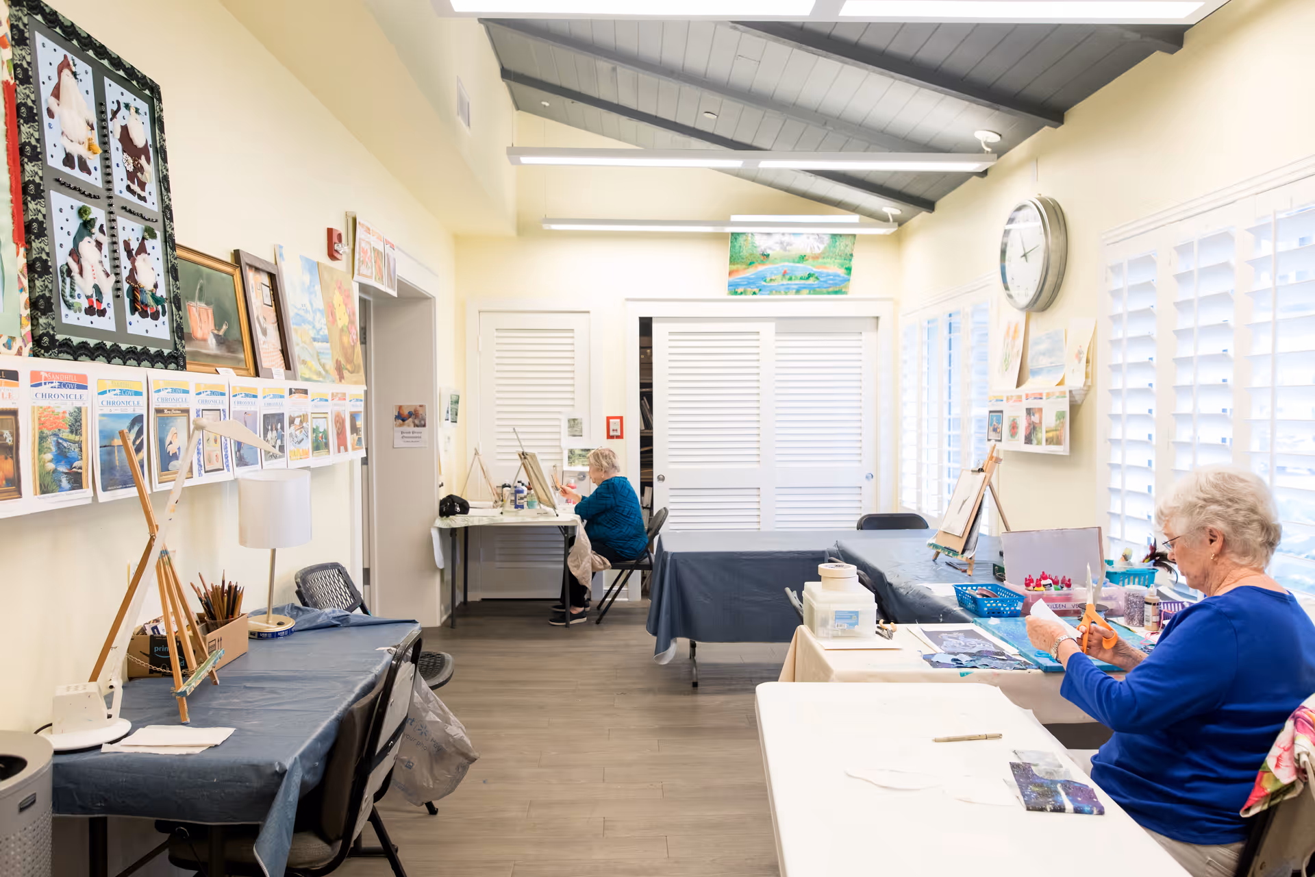 A bright, well-lit room with tables covered in blue and white tablecloths, where two elderly women are engaged in arts and crafts activities. The walls are decorated with various paintings, artwork, and a large clock. The room has large windows with white shutters allowing natural light to enter.