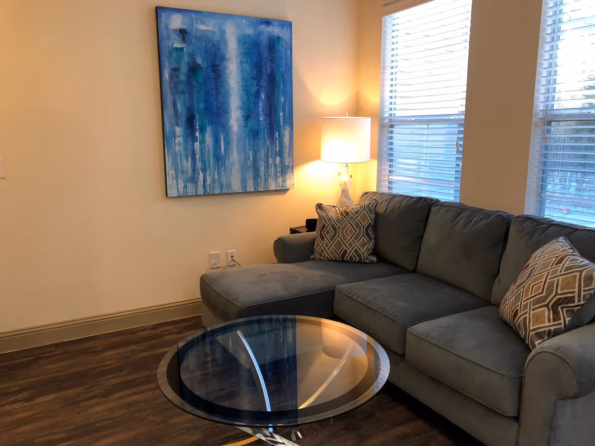 A cozy living room area with a gray sectional sofa adorned with patterned throw pillows, a round glass coffee table, a lit table lamp on a side table, and a large abstract blue painting on the wall. Two windows with white blinds allow natural light into the room.
