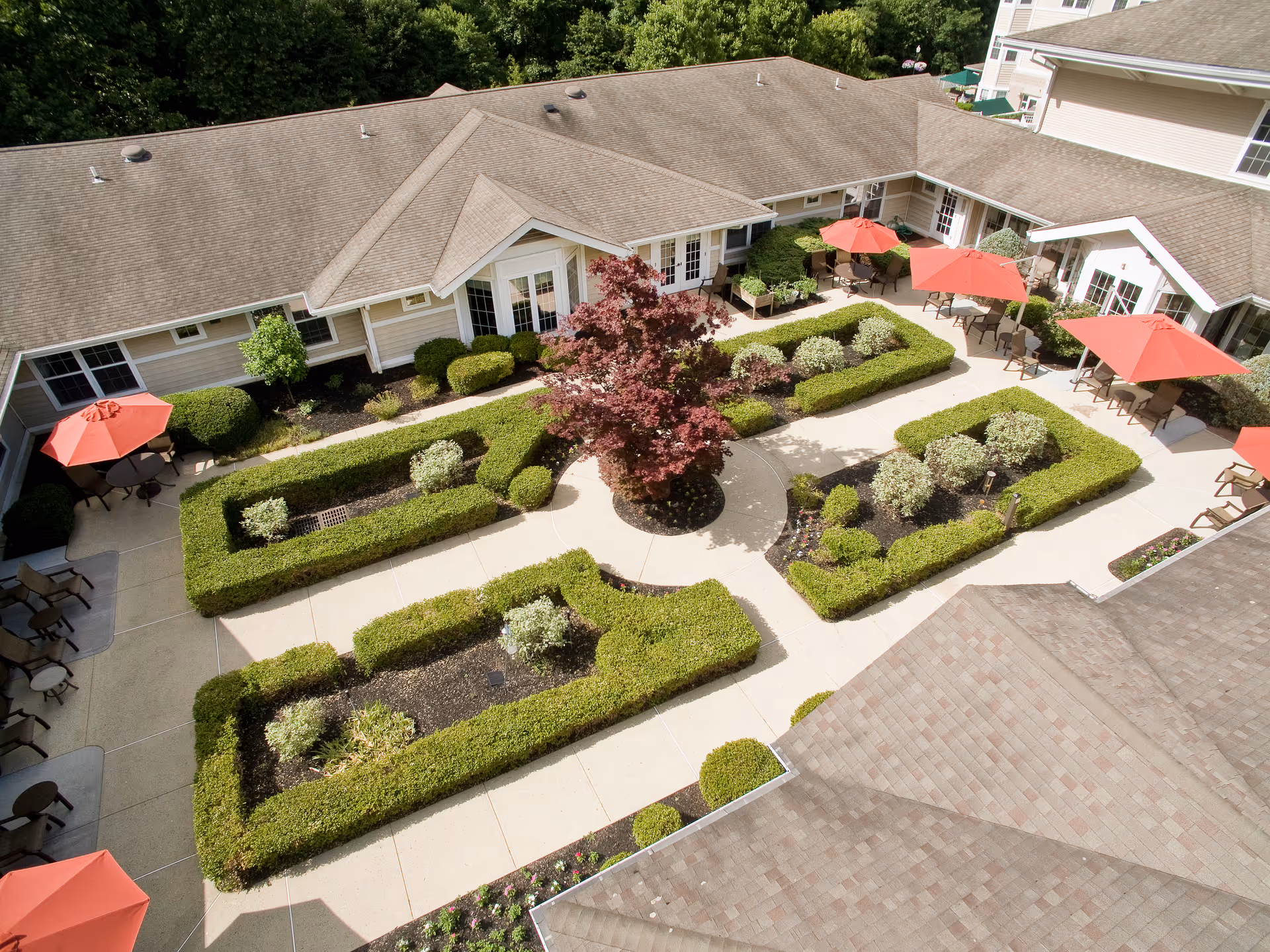 Aerial view of a landscaped central courtyard with trimmed hedges, walkways, red patio umbrellas and surrounding senior living building.