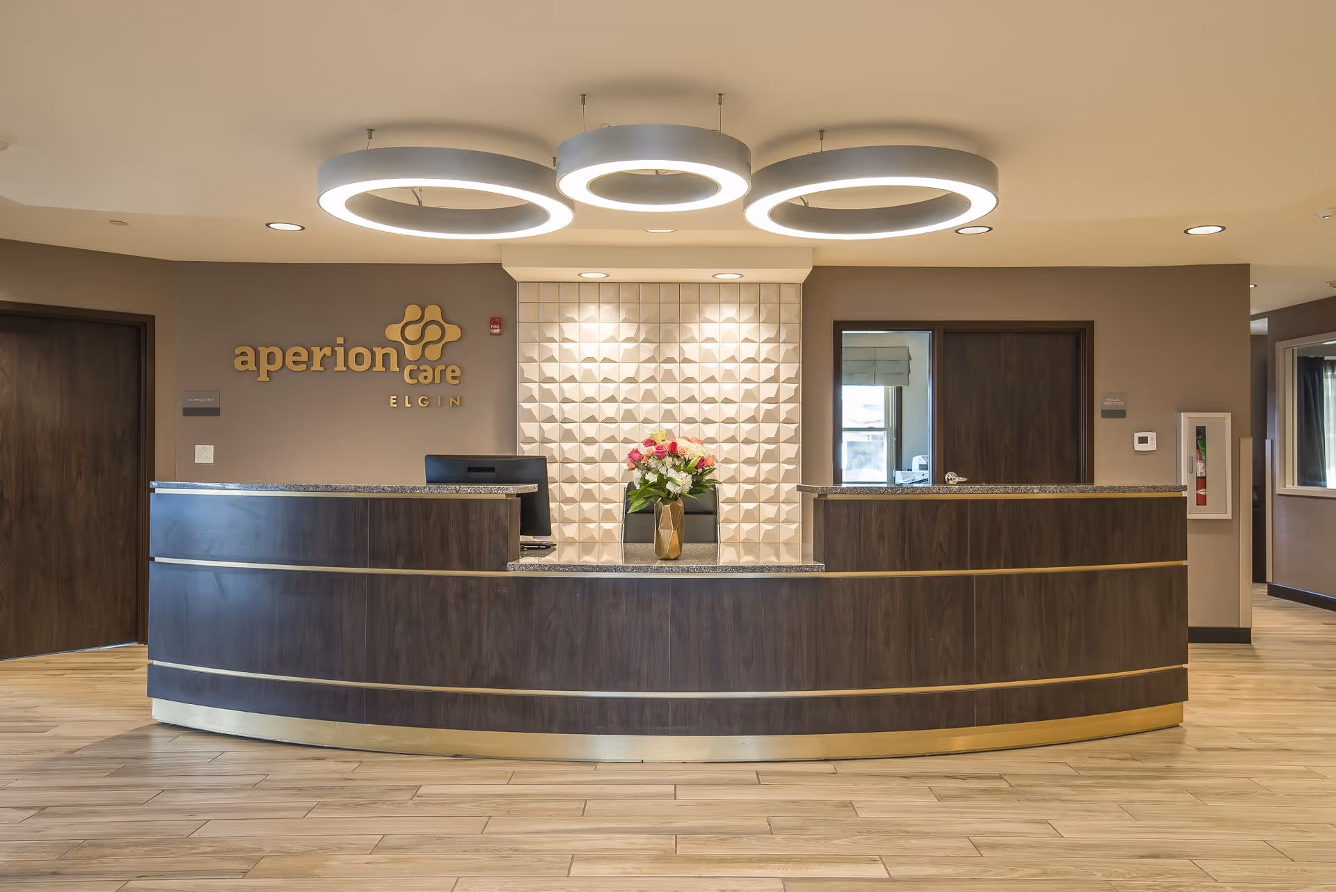 Reception area of Aperion Care Elgin featuring a curved dark wood front desk with a granite countertop, a vase with flowers on the desk, modern circular ceiling lights, and a textured white wall panel behind the desk. The facility's name and logo are displayed on the wall to the left of the desk.