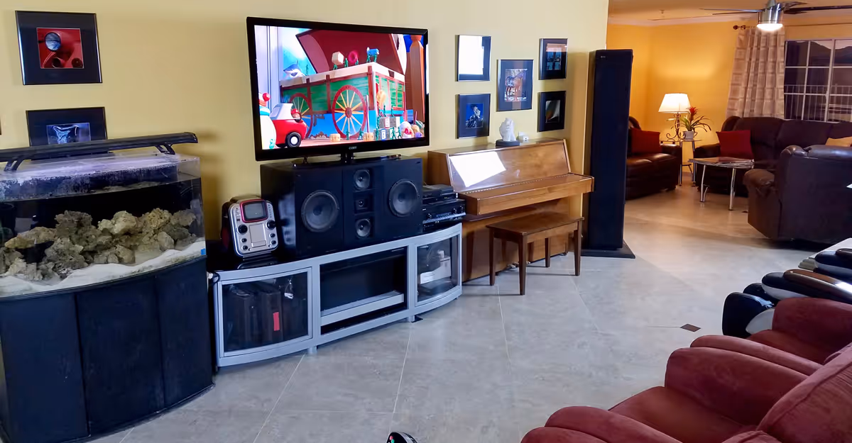 Living room area with a wall-mounted TV above a media console and speakers, a large aquarium, a piano, and several recliners and sofas.