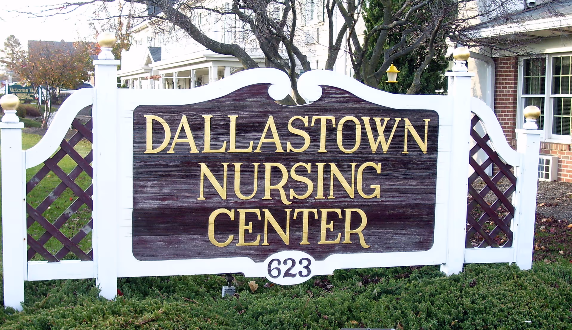 A wooden sign with white posts and lattice sides displaying the text 'Dallastown Nursing Center' in gold letters and the number '623' below, set in a landscaped area with greenery and a building partially visible in the background.