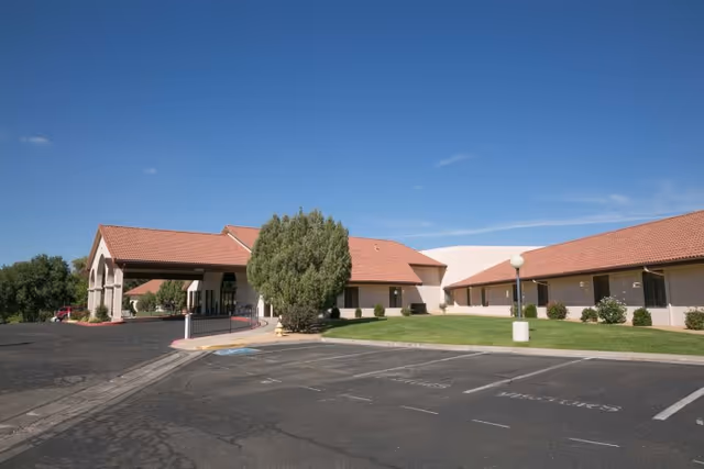 Single-story red-tile roof health and rehabilitation building with a covered entrance, parking lot, and landscaped lawn under a clear blue sky.