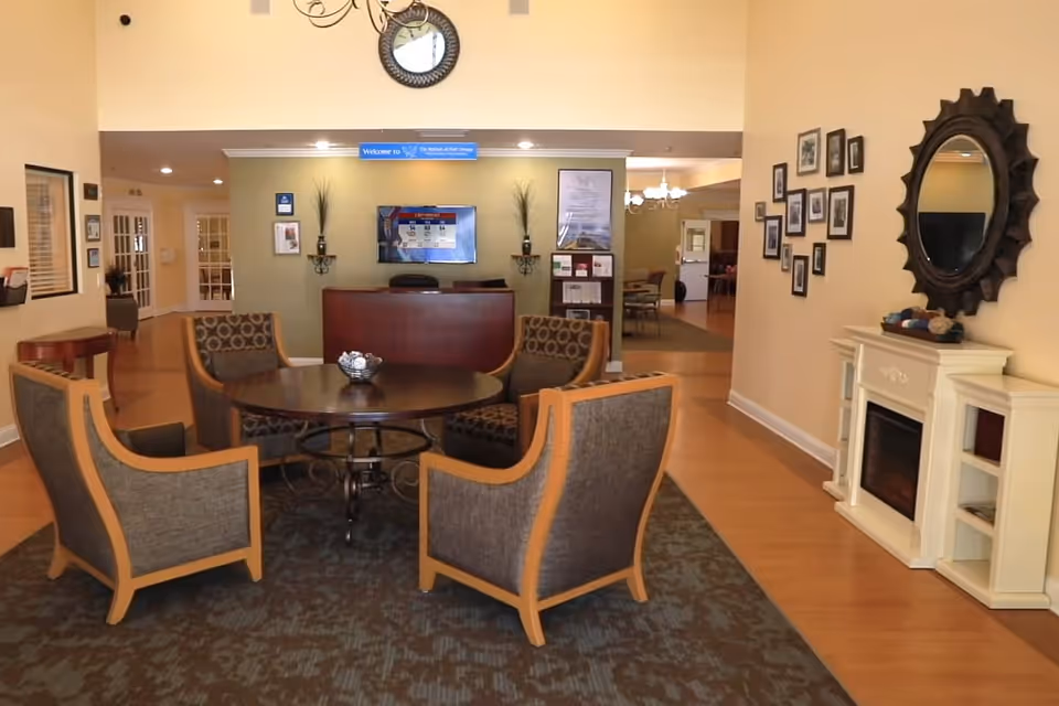 A senior living facility reception/lounge with a round table surrounded by four armchairs, a front desk in the background, wall mirror and a decorative fireplace.