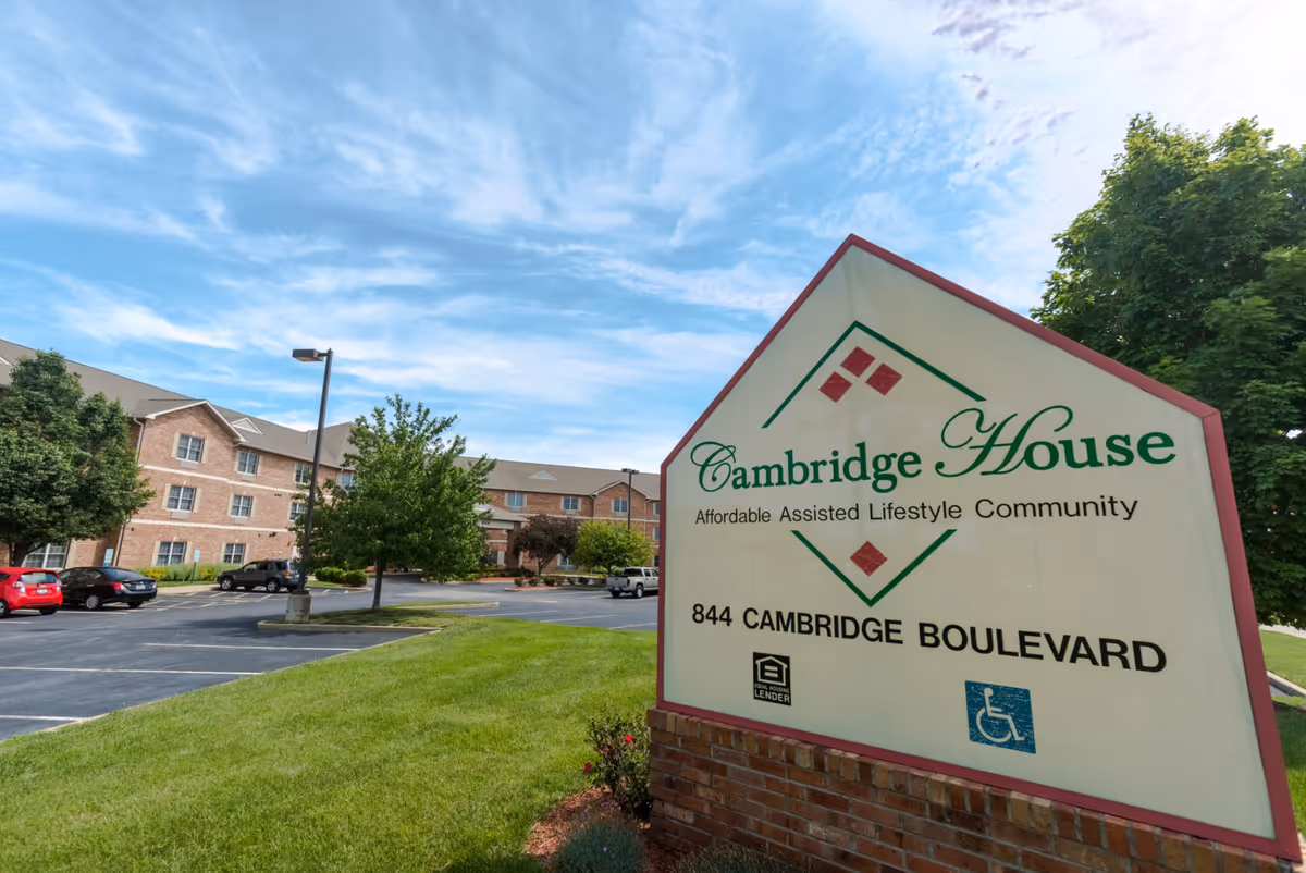 Outdoor view of Cambridge House of Ofallon, showing a large sign with the facility name, address, and accessibility symbols in front of a brick building with multiple windows, surrounded by trees and a parking lot under a partly cloudy sky.