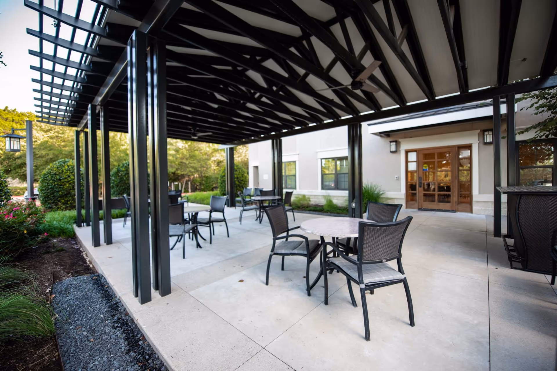 Outdoor covered patio area at Belmont Village Senior Living West Lake Hills with several round tables and black chairs arranged on a concrete floor. The patio has a black metal pergola overhead and is surrounded by greenery and bushes. The building entrance with glass doors is visible in the background.