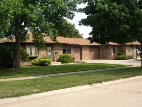 Single-story brick senior living facility exterior with manicured lawn and trees along the street.