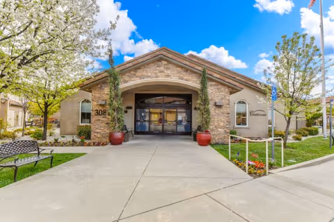 Front entrance of a clubhouse-style building with a stone facade, arched entry, potted plants, and blooming trees under a blue sky.