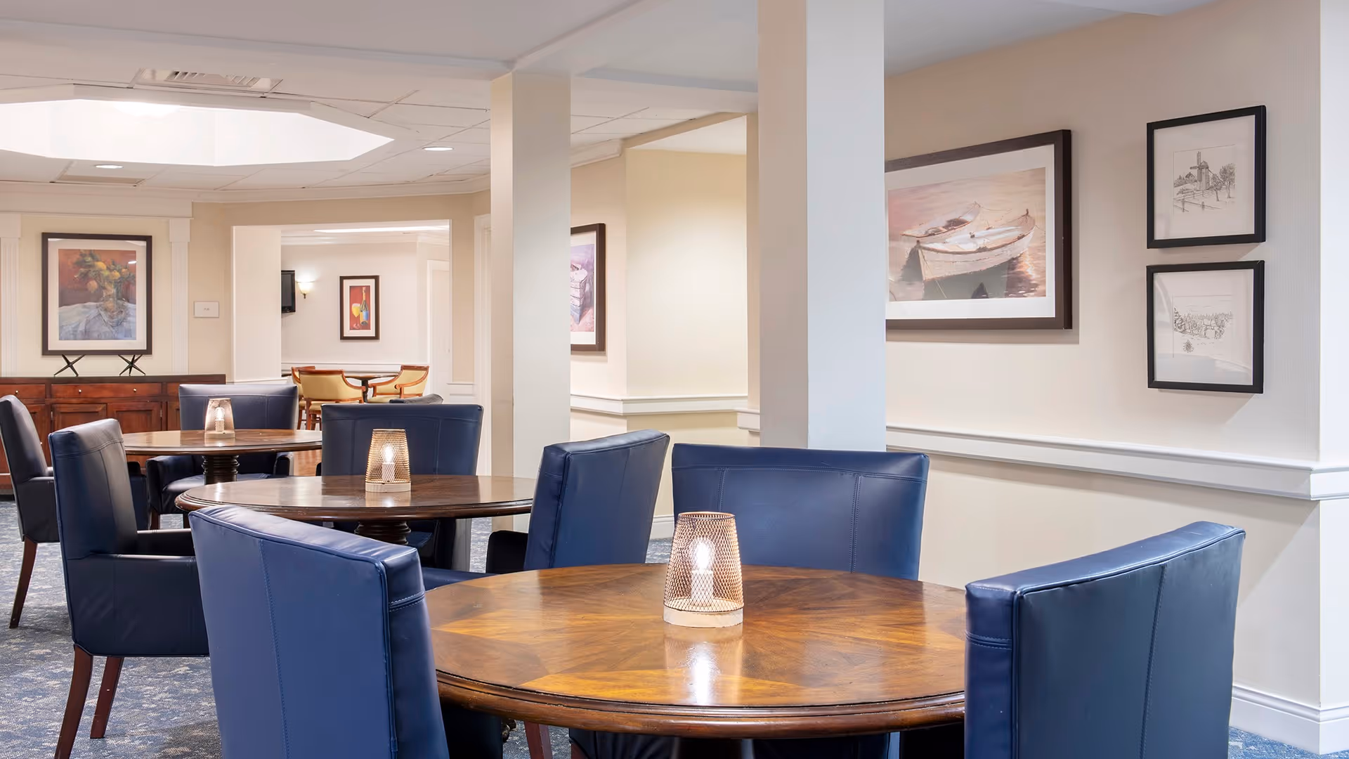 A cozy dining area with round wooden tables and blue upholstered chairs. Each table has a candle-like light centerpiece. The walls are decorated with framed artwork, including paintings and sketches. The room has soft lighting and a calm, inviting atmosphere.