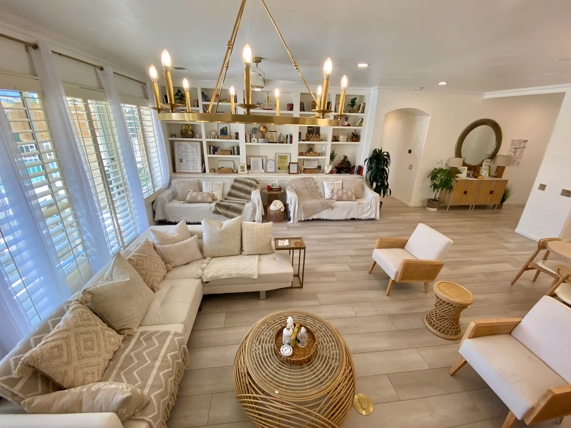 A bright and spacious living room with large windows covered by white sheer curtains. The room features a beige sectional sofa with multiple cushions, two armchairs with wooden frames and light upholstery, and a round wicker coffee table with decorative items on top. A modern gold chandelier hangs from the ceiling. In the background, there are two white sofas with throws and pillows, a built-in white bookshelf filled with books and decorative items, and a wooden sideboard with plants and a round mirror above it. The floor is light wood, and the walls are painted white.