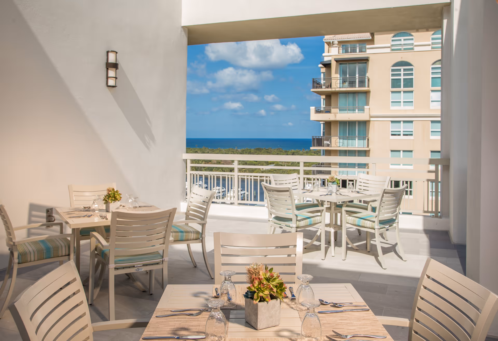 Outdoor dining area with several tables and chairs arranged on a balcony or terrace. Each table is set with placemats, glassware, and cutlery, and decorated with small potted plants. The area overlooks a scenic view of the ocean, greenery, and a nearby multi-story building under a partly cloudy sky.