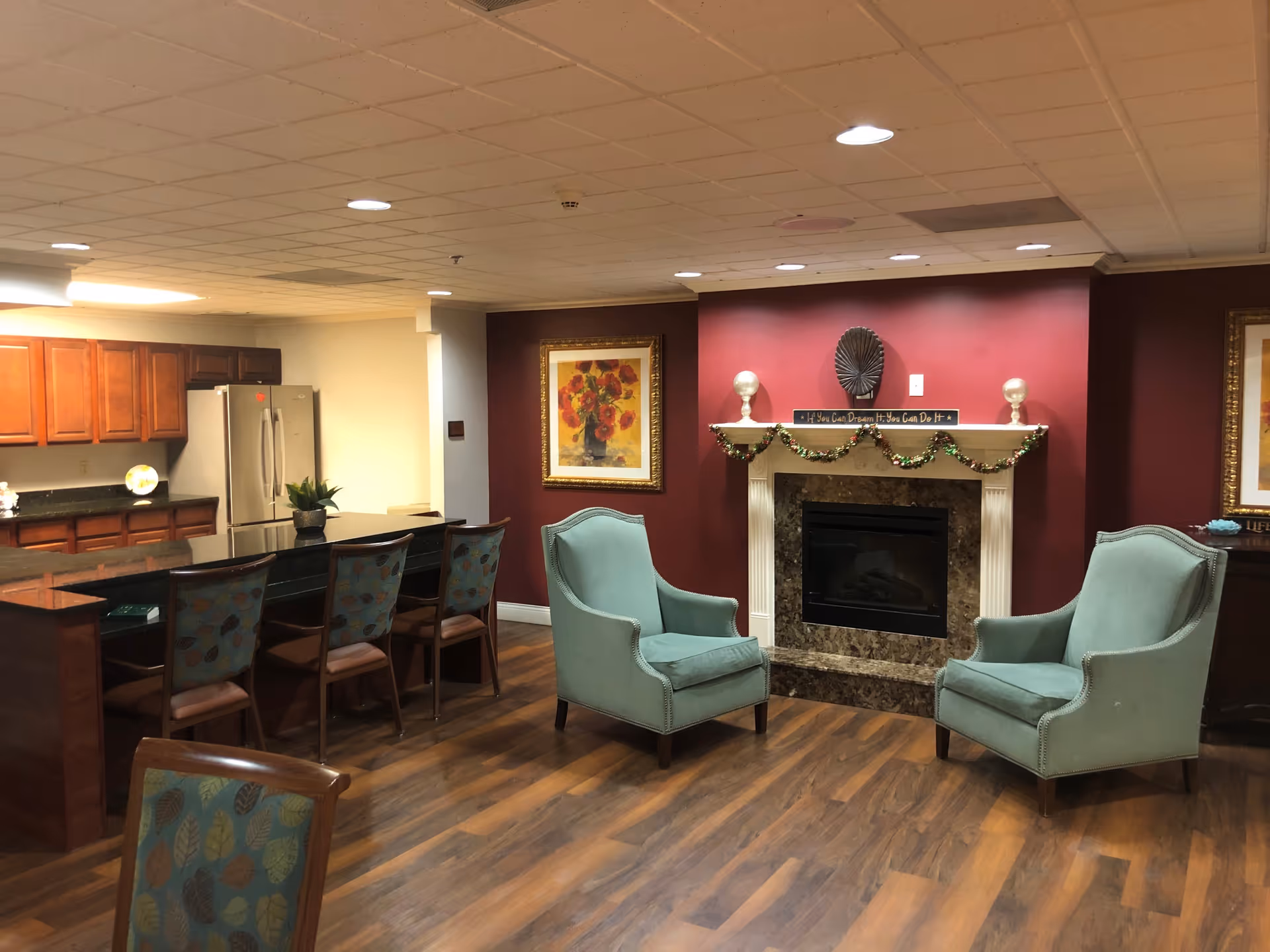 Interior view of a senior living facility common area featuring a kitchen with wooden cabinets and a stainless steel refrigerator, a granite countertop with four chairs, two teal upholstered armchairs facing a fireplace decorated with a garland and a sign that reads 'If You Can Dream It, You Can Do It', and framed floral artwork on the walls.