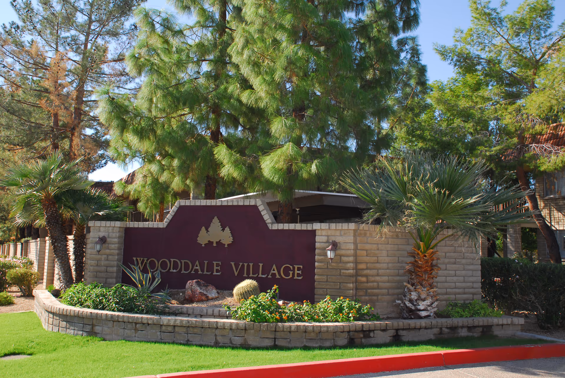 Entrance sign for Wooddale Village Retirement facility surrounded by landscaped greenery including palm trees, bushes, and other plants, with a brick wall and a clear blue sky in the background.