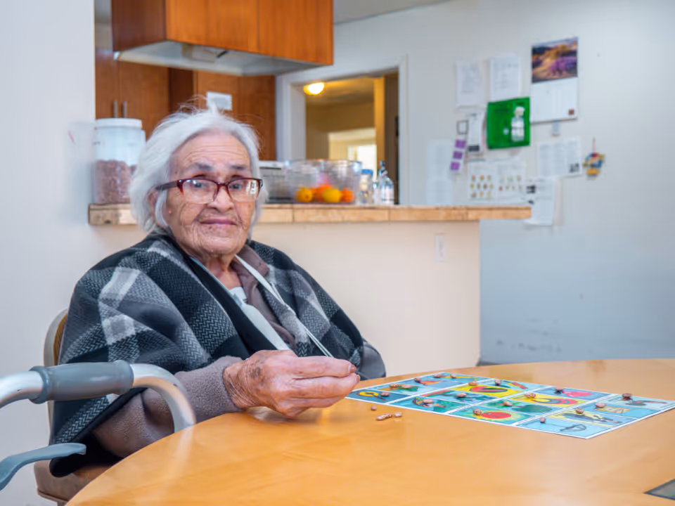 An elderly woman with white hair and glasses sits at a wooden table in a kitchen or dining area. She is wearing a plaid shawl and holding small objects in her hand, with a colorful game board on the table in front of her. The background shows kitchen cabinets, a counter with fruit bowls, and a wall with various papers and a calendar.