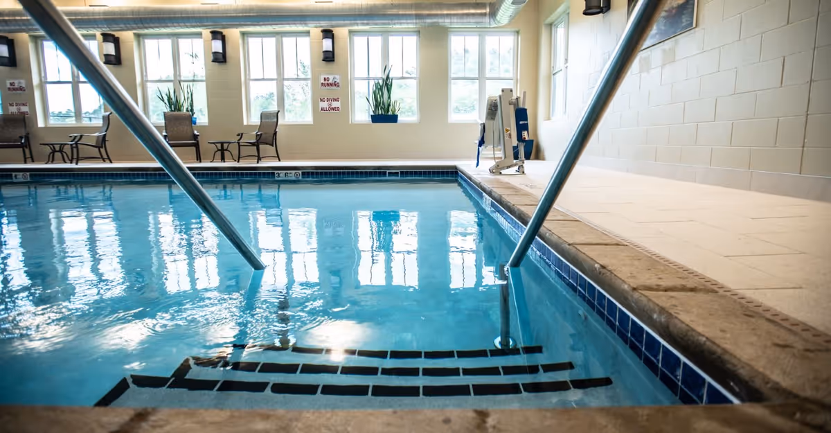 Indoor swimming pool with metal handrails and steps, windows and seating along the far wall.