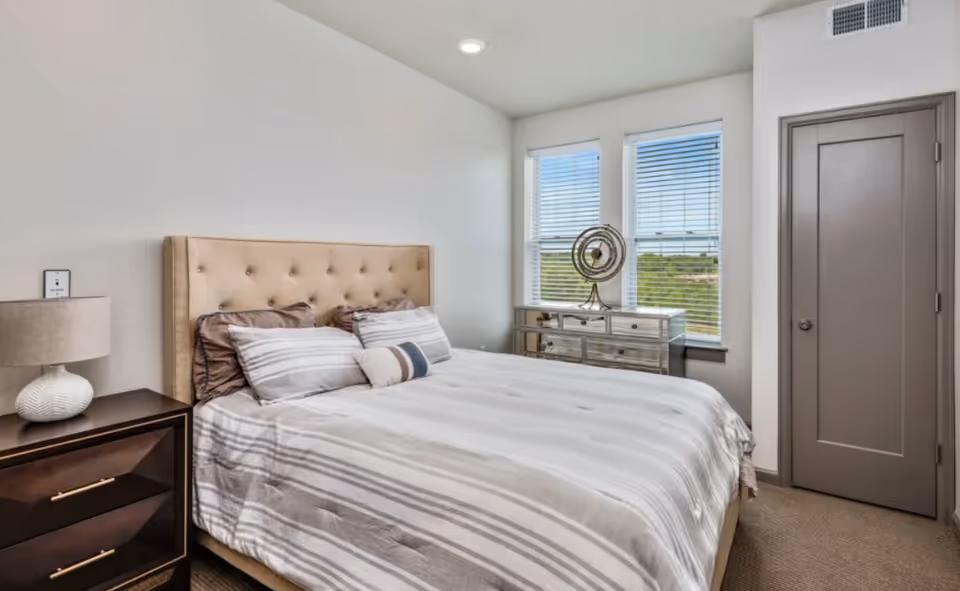 A bedroom with a beige tufted headboard bed dressed in striped bedding and multiple pillows. To the left of the bed is a dark wooden nightstand with a white textured lamp. On the right side of the room, there is a mirrored dresser with decorative items on top, positioned in front of two windows with white blinds. A closed gray door is visible on the right wall.