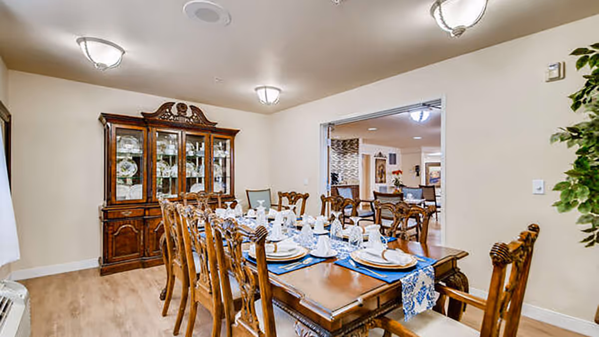 A formal dining room with a long wooden dining table set for a meal with white plates, glasses, and napkins on blue placemats. The room features wooden chairs with intricate carvings, a large wooden china cabinet filled with dishes, and light-colored walls and flooring. There is an open doorway leading to another room with additional seating and decor.