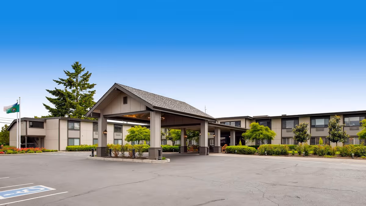 Exterior view of Cascade Inn Assisted Living Community showing a covered entrance with a peaked roof, surrounded by a parking lot and landscaped greenery including trees and bushes under a clear blue sky.